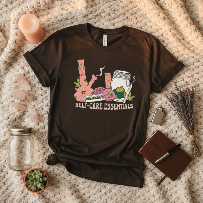 A dark brown short-sleeve t-shirt with a colorful graphic on the front showing self-care items like a pink bong, a pill bottle, a jar, and a green leaf, with the text 'SELF-CARE ESSENTIALS' underneath.