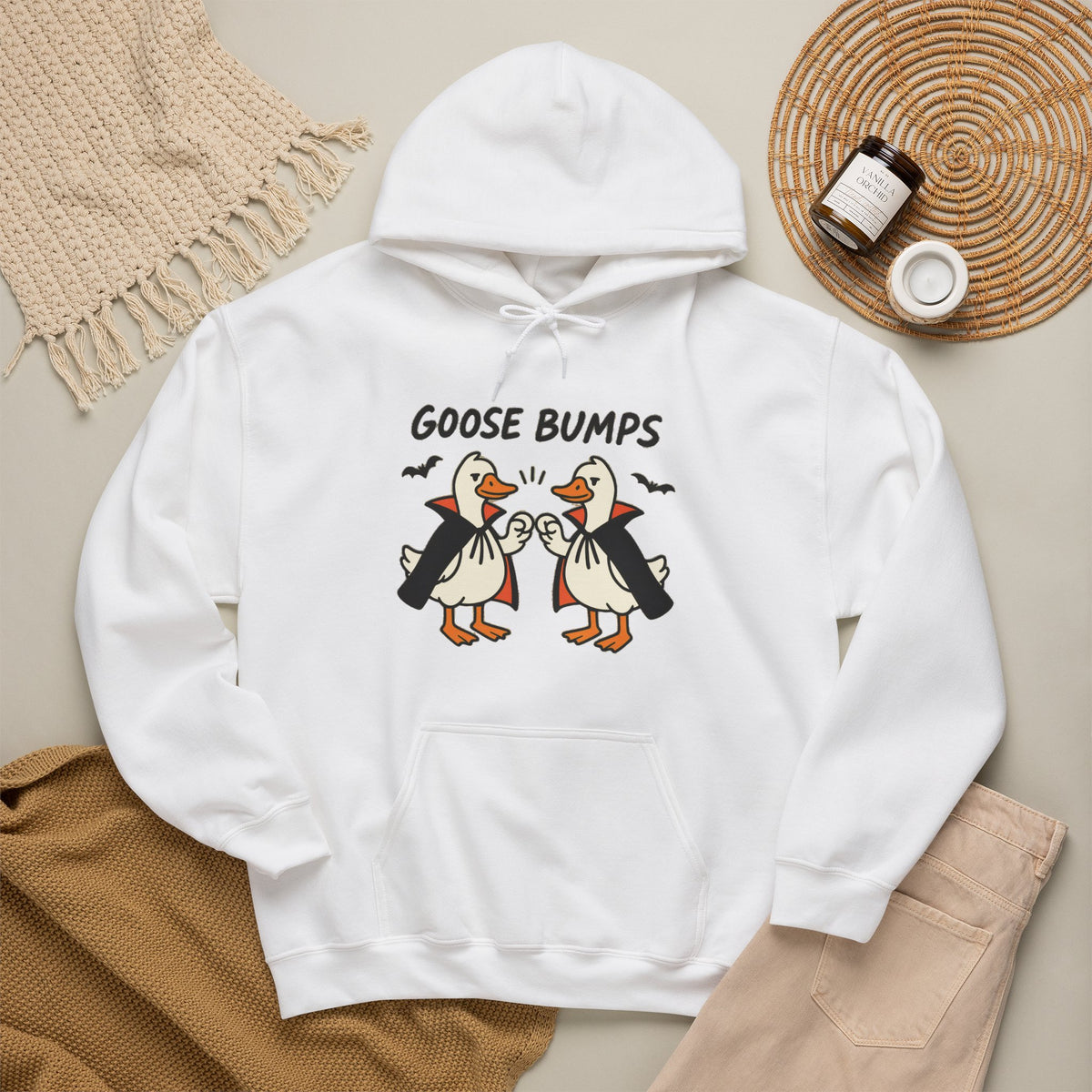 A white pullover hoodie laid flat on a beige surface, featuring a graphic of two cartoon geese in vampire outfits with the text 'GOOSE BUMPS' above them. The hoodie is styled with a tan knit blanket, beige pants, and a small jar on a woven mat.