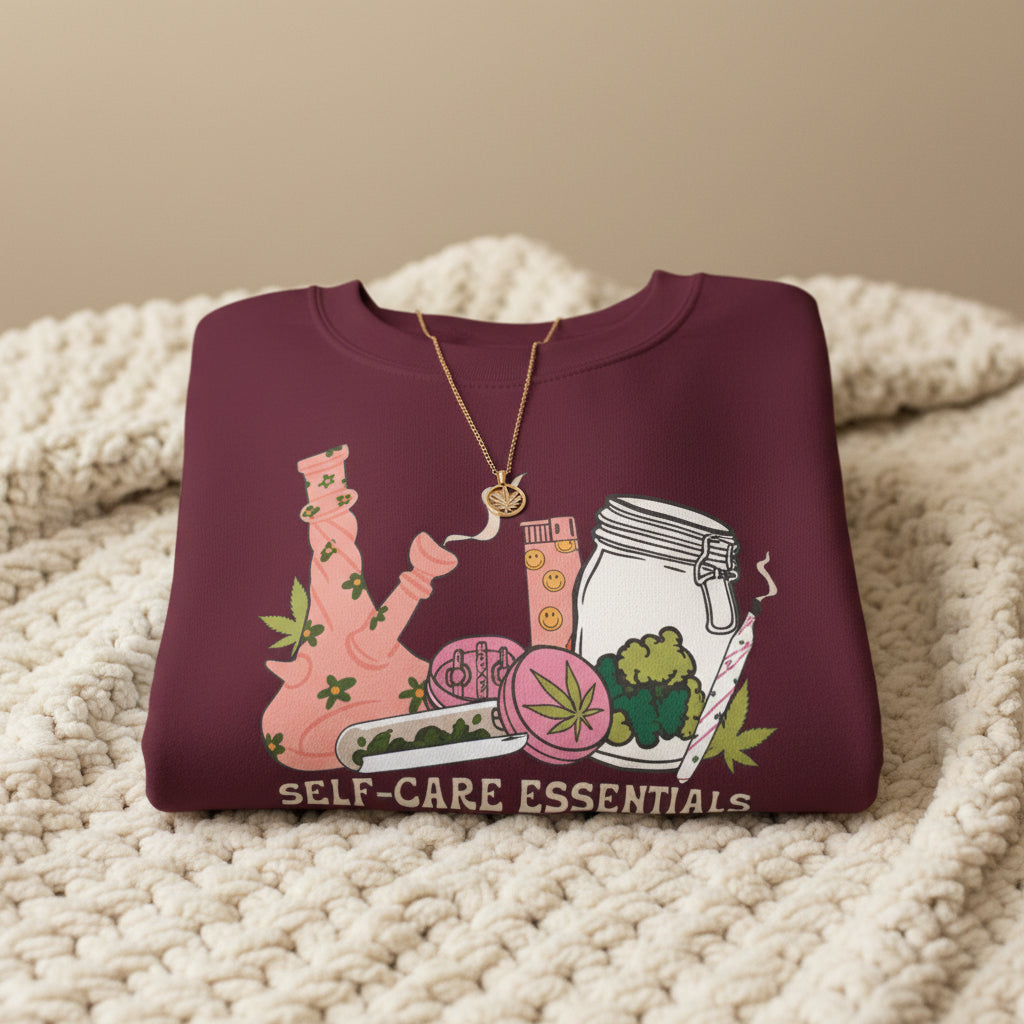 A maroon crewneck sweatshirt featuring a colorful graphic of self-care and healing elements, including a pink bong, a jar, a cup, a flower, and a rolling joint, with the text 'SELF-CARE ESSENTIALS' printed below.