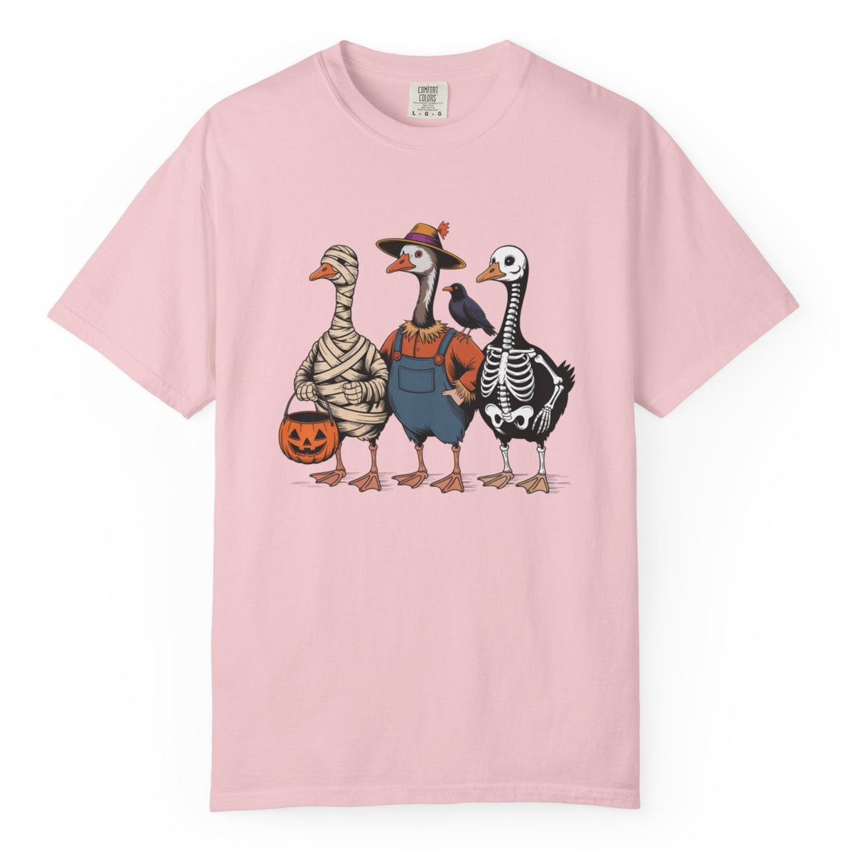 A light pink short-sleeve crewneck t-shirt featuring a graphic print of three geese dressed in Halloween costumes (mummy, skeleton, and cowboy), set against a plain white background.
