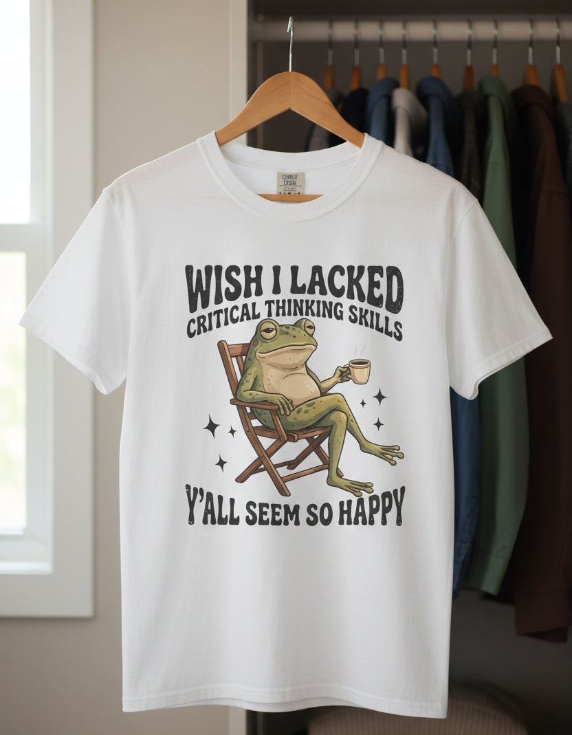 A white short-sleeve crewneck t-shirt featuring a graphic of a green frog sitting in a wooden director's chair and holding a coffee mug, with humorous text above and below the illustration.