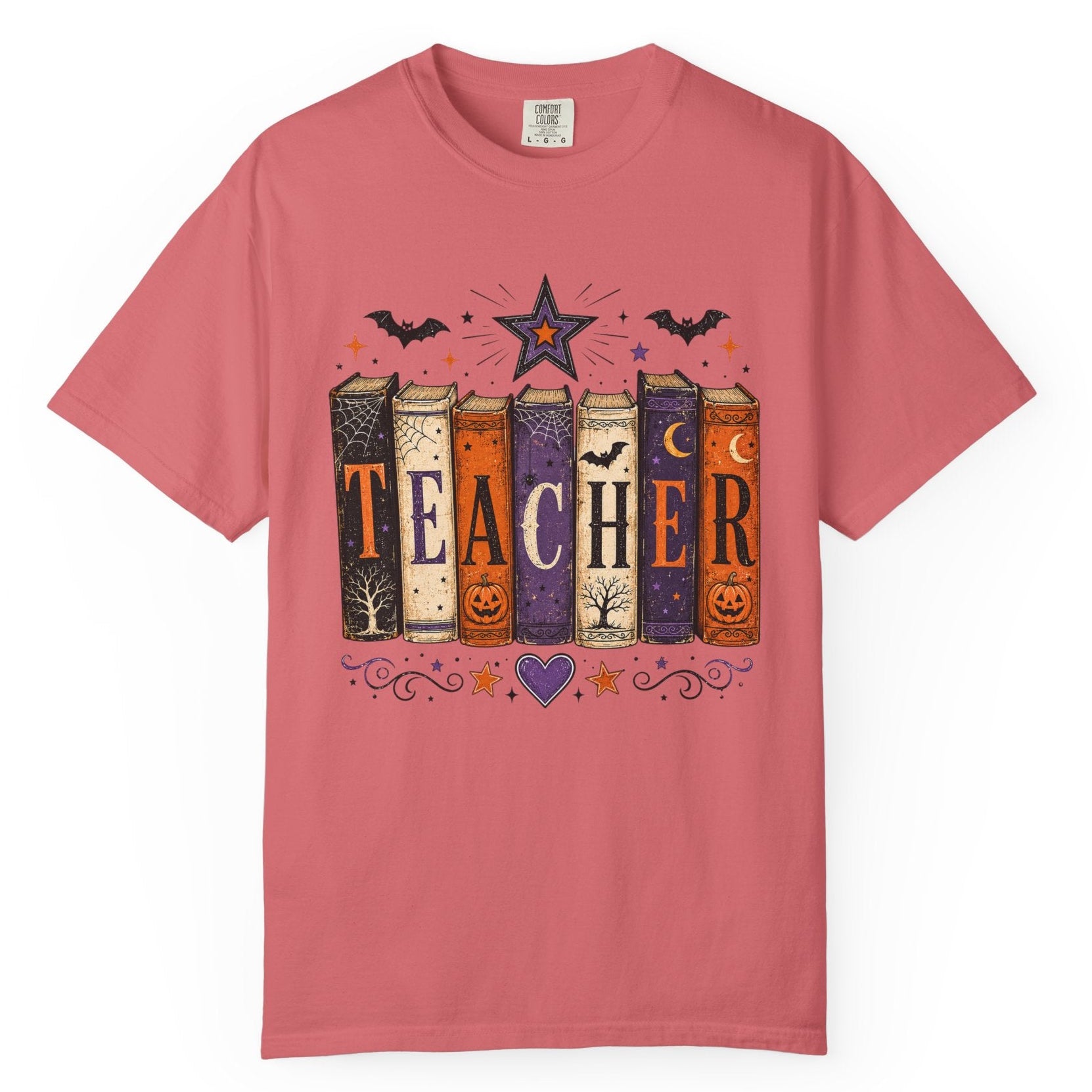 A pink short-sleeve crewneck t-shirt featuring a Halloween-themed graphic of six books spelling 'TEACHER' with spooky illustrations like bats, a star, and jack-o'-lanterns.