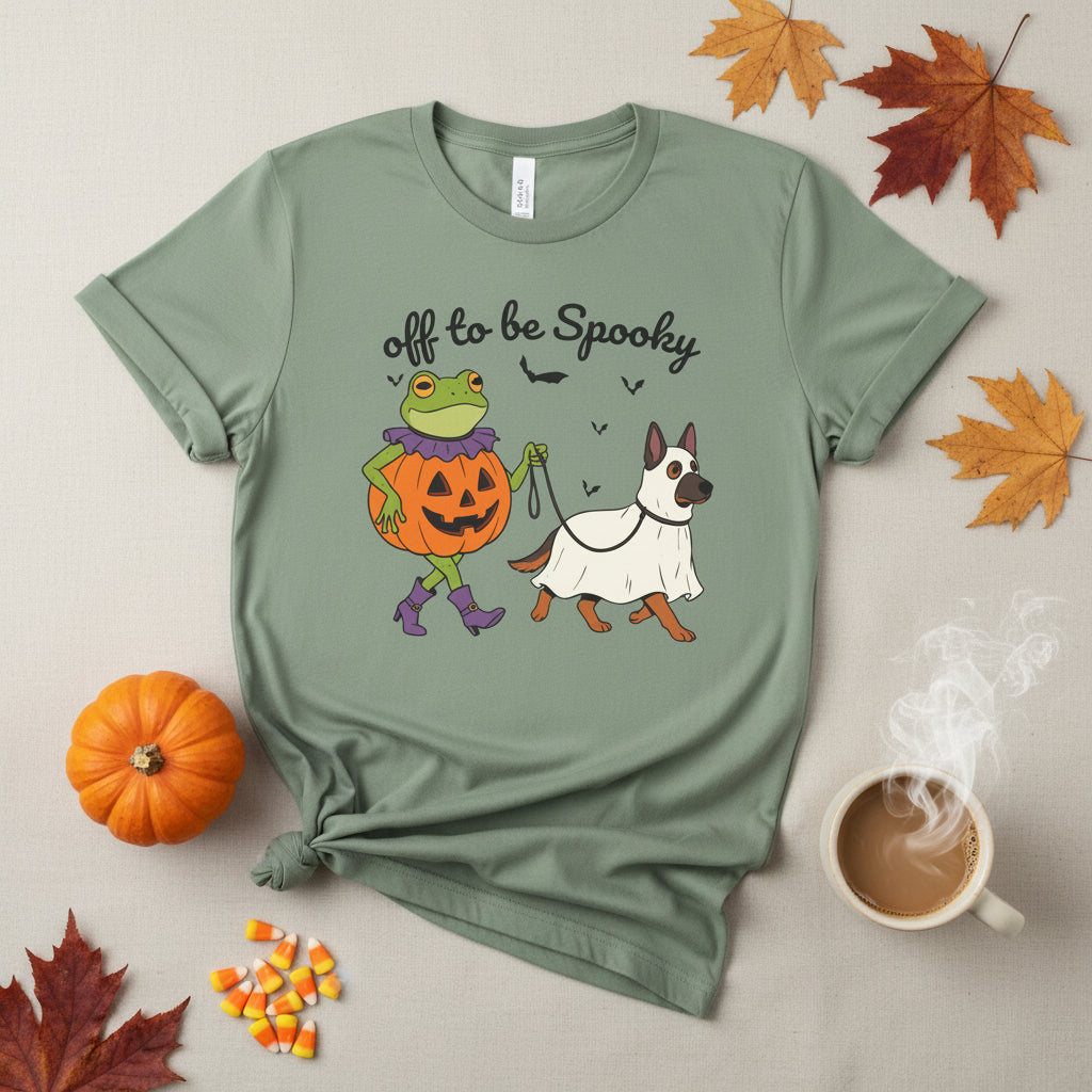A sage green short-sleeve t-shirt with a Halloween graphic of a frog in a pumpkin costume and a dog in a white ghost costume, with the text 'off to be Spooky' above them.