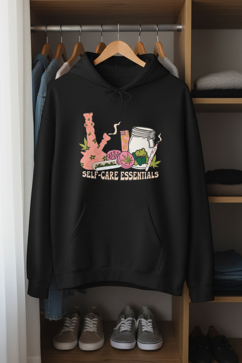 A black pullover hoodie with a colorful graphic on the chest depicting a pink bong, a pink drink, a rolling machine, a jar, and the words 'SELF-CARE ESSENTIALS' in cream text, shown against a plain white background.