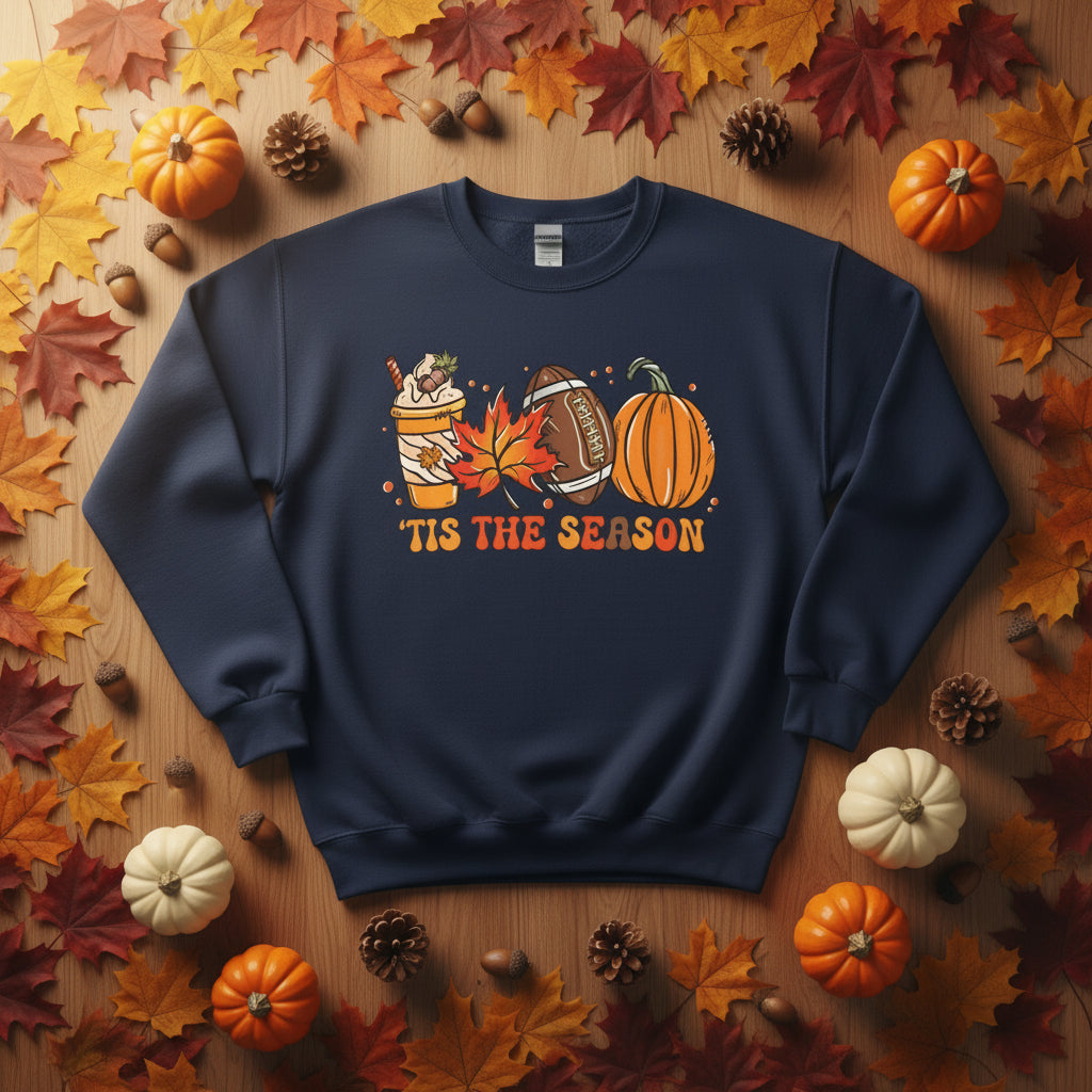 A navy blue crewneck sweatshirt featuring a festive autumn graphic with a pumpkin, football, hot chocolate cup, falling leaves, and the text 'TIS THE SEASON' in orange, 