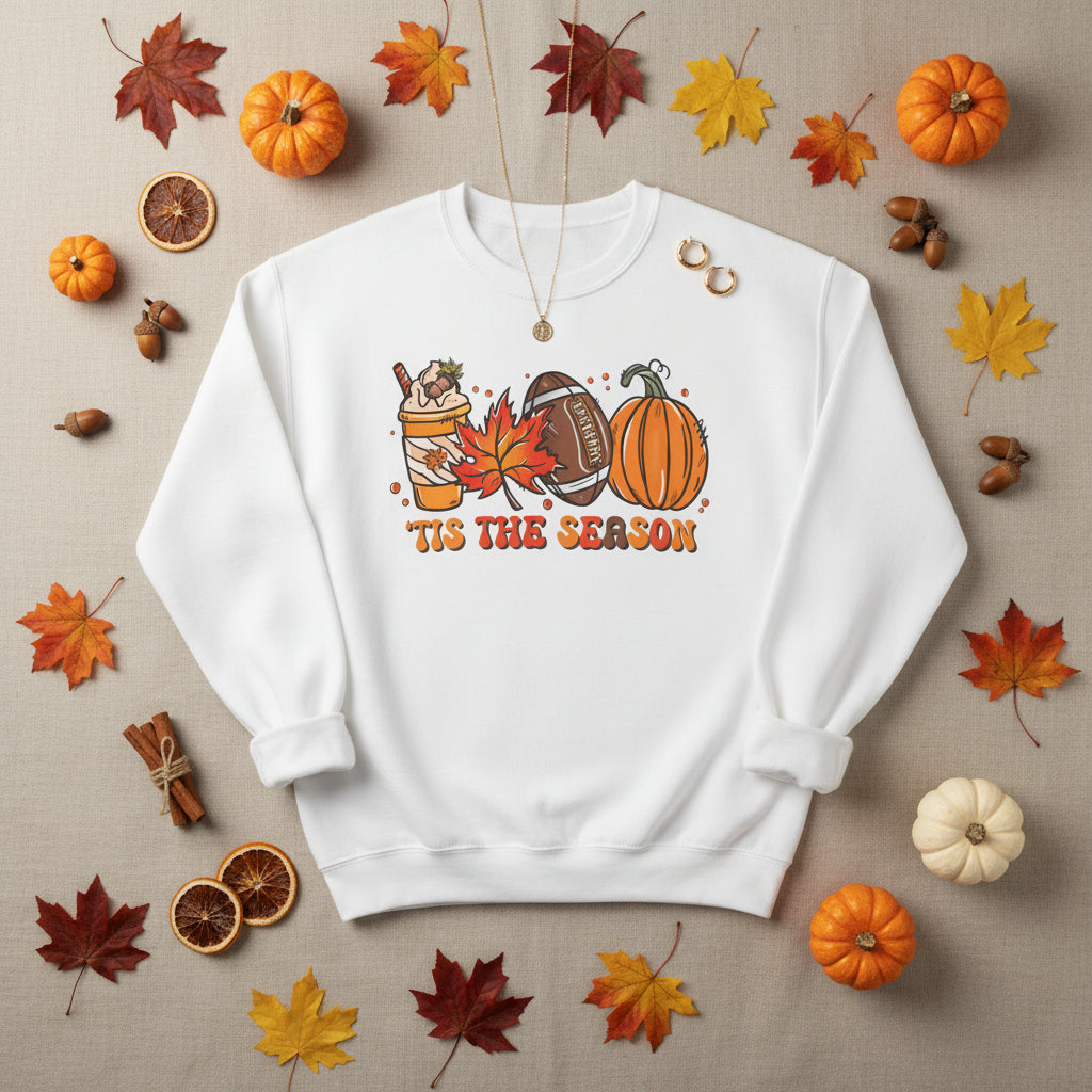 An white crewneck sweatshirt featuring a colorful autumn-themed graphic with a drink, maple leaf, football, pumpkin, and the text 'TIS THE SEASON'.