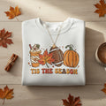 A light heather gray crewneck sweatshirt featuring a colorful autumn-themed graphic with a hot chocolate cup, maple leaf, football, pumpkin, and the text 'TIS THE SEASON' in orange.