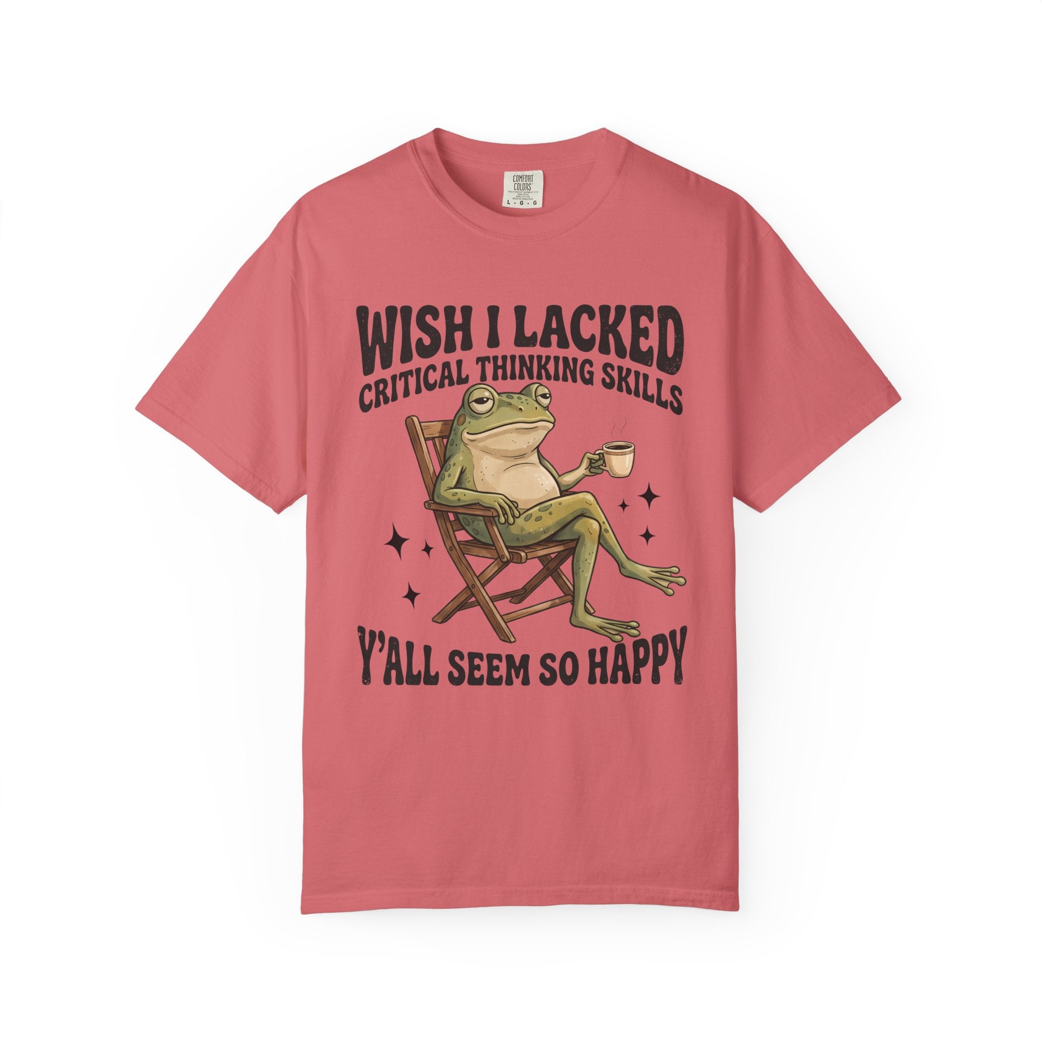 A pink short-sleeve crewneck t-shirt featuring a graphic of a green frog sitting in a wooden chair holding a mug, with the text 'WISH I LACKED CRITICAL THINKING SKILLS' above and 'Y'ALL SEEM SO HAPPY' below.