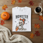 A white short-sleeve crewneck t-shirt featuring a graphic of a skeleton woman with a black bow and orange hair accessory, holding an orange drink, with the word 'MOMSTER' printed above in a distressed black font.