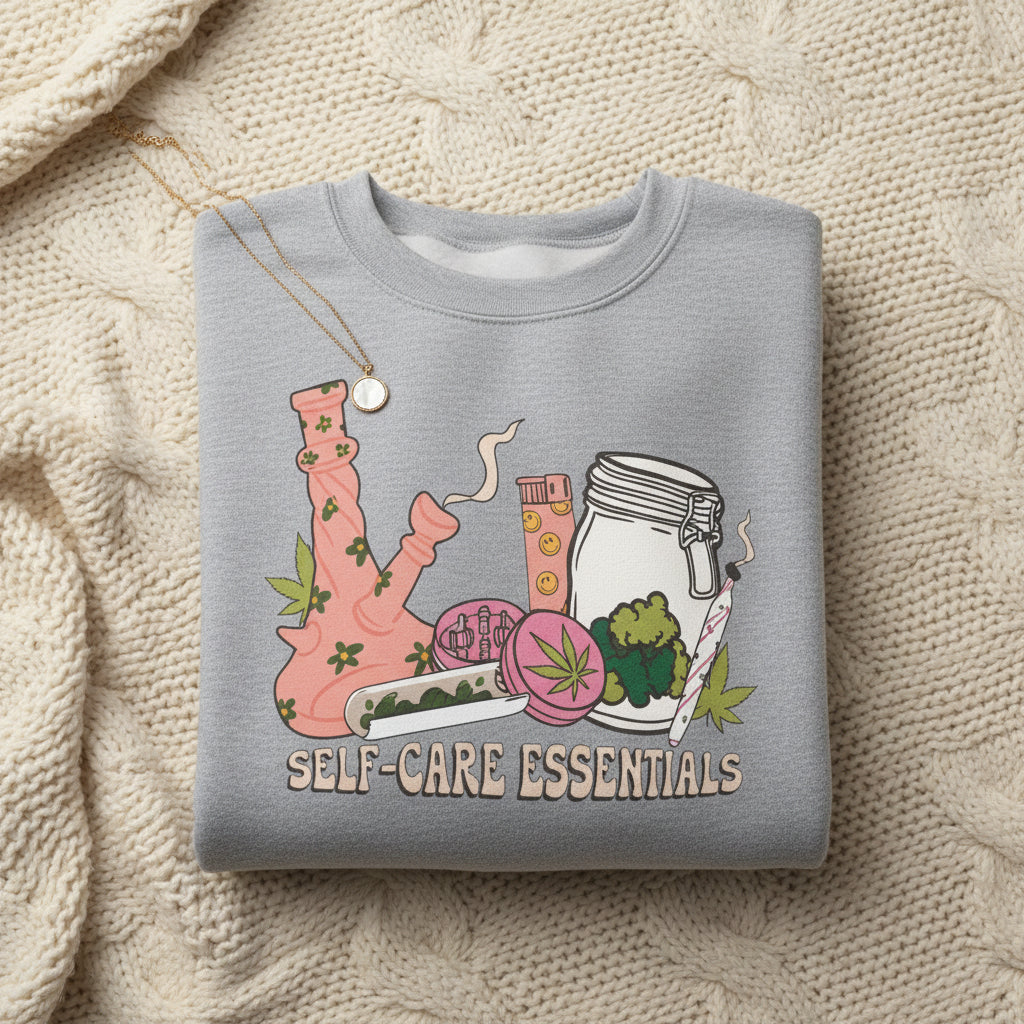 A heather gray crewneck sweatshirt featuring a colorful graphic of a pink bong, a glass jar, a pink cup, and a purple flower with the text 'SELF-CARE ESSENTIALS' printed below.