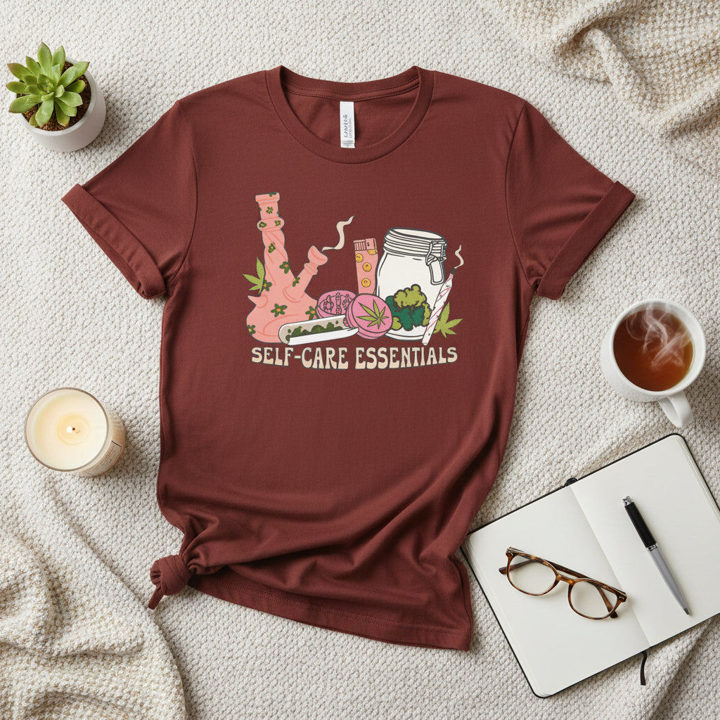 A rust-brown short-sleeve crewneck t-shirt featuring a colorful graphic of self-care items arranged to spell out 'K' with the text 'SELF-CARE ESSENTIALS' underneath, displayed on a solid white background.