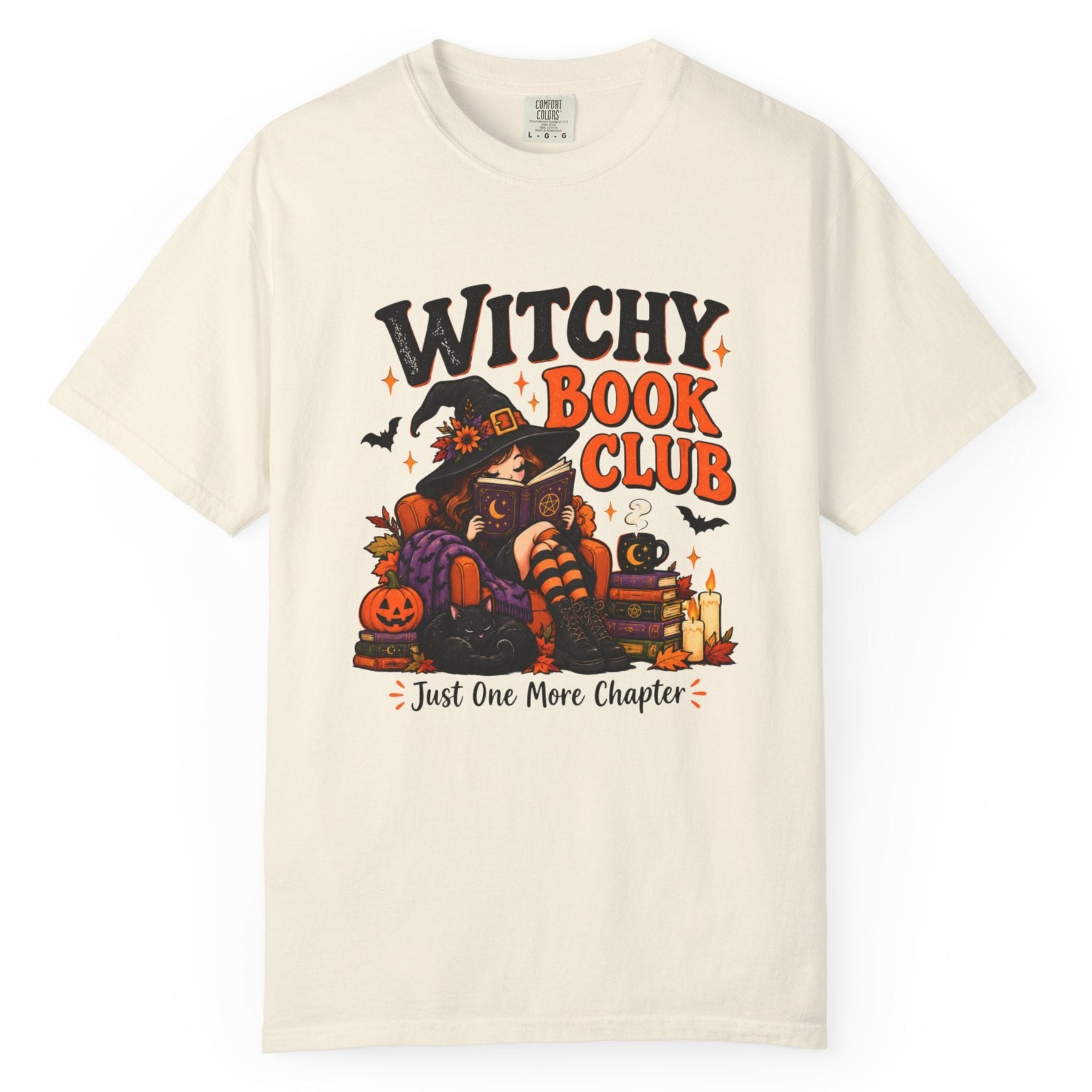 An off-white short-sleeve t-shirt with a colorful graphic on the front that reads 'WITCHY BOOK CLUB' and 'Just One More Chapter' above a Halloween-themed illustration of a witch, pumpkin, and cat.