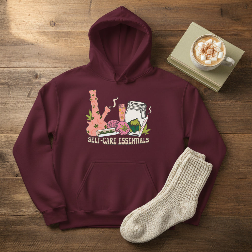 A maroon pullover hoodie with a colorful graphic on the chest depicting self-care elements like a pink bong, a cup, a jar, and a rolling machine, with the text 'SELF-CARE ESSENTIALS' printed below.