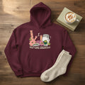 A maroon pullover hoodie with a colorful graphic on the chest depicting self-care elements like a pink bong, a cup, a jar, and a rolling machine, with the text 'SELF-CARE ESSENTIALS' printed below.