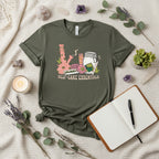 An army short-sleeve t-shirt with a colorful graphic on the front showing a pink bong, a glass jar, a pink cup, and a green leaf, with the text 'SELF-CARE ESSENTIALS' printed below.