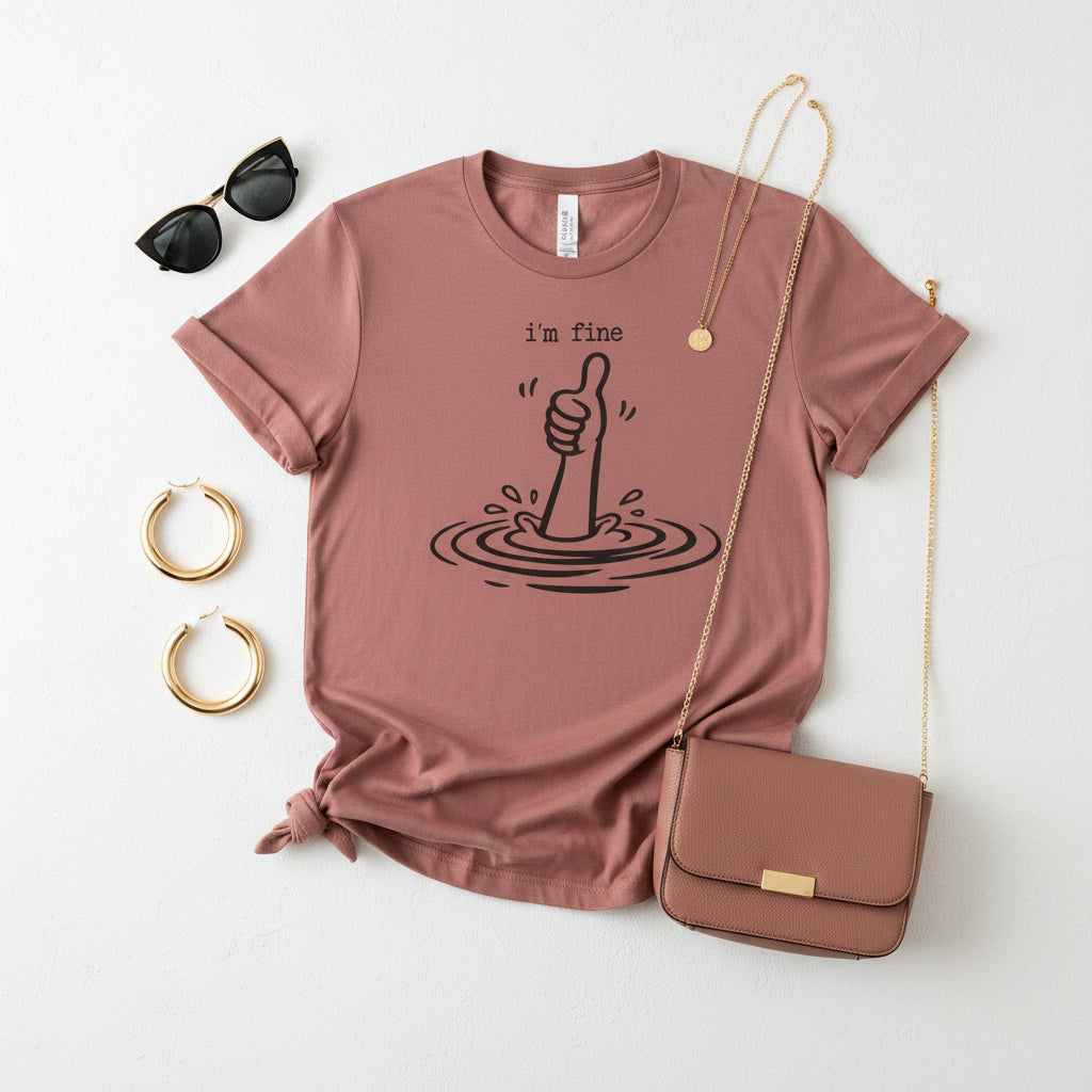 A pink short-sleeve crewneck t-shirt featuring a black graphic of a hand giving a thumbs-up with ripples and the text 'i'm fine' printed on the chest.