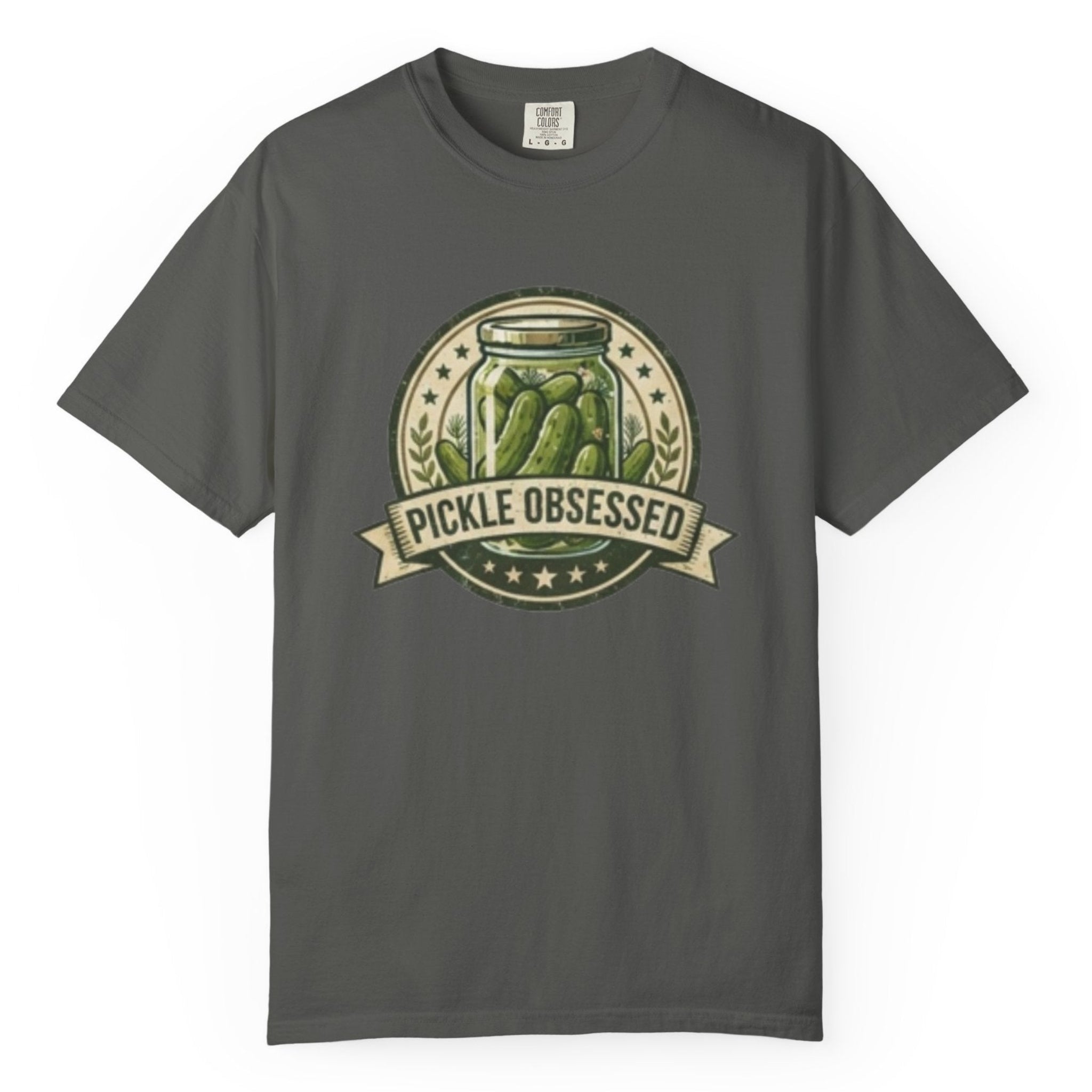 A pepper short-sleeve t-shirt hanging on a wooden hanger against a light wood pegboard wall. The shirt features a circular graphic print with a jar of pickled green onions and the words 'PICKLE OBSESSED' in a banner.