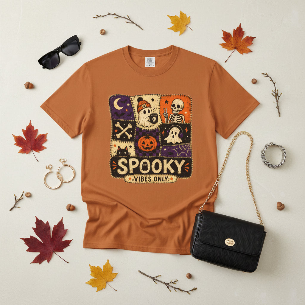 A rust-orange crewneck t-shirt featuring a central square graphic with a patchwork Halloween theme, including a skeleton, moon, jack-o'-lantern, ghosts, and the text 'SPOOKY VIBES ONLY'.