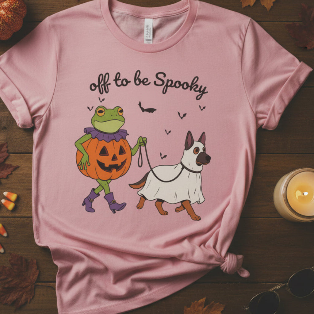 A light pink short-sleeve crewneck t-shirt featuring a Halloween graphic of a green frog dressed as a pumpkin and a white dog in a ghost costume, with the text 'off to be Spooky' above them.