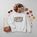A light heather gray crewneck sweatshirt featuring the text 'COFFEE WEATHER' printed on the chest. The letters in 'COFFEE' are filled with a brown and beige checkerboard pattern, and both words are outlined in brown.