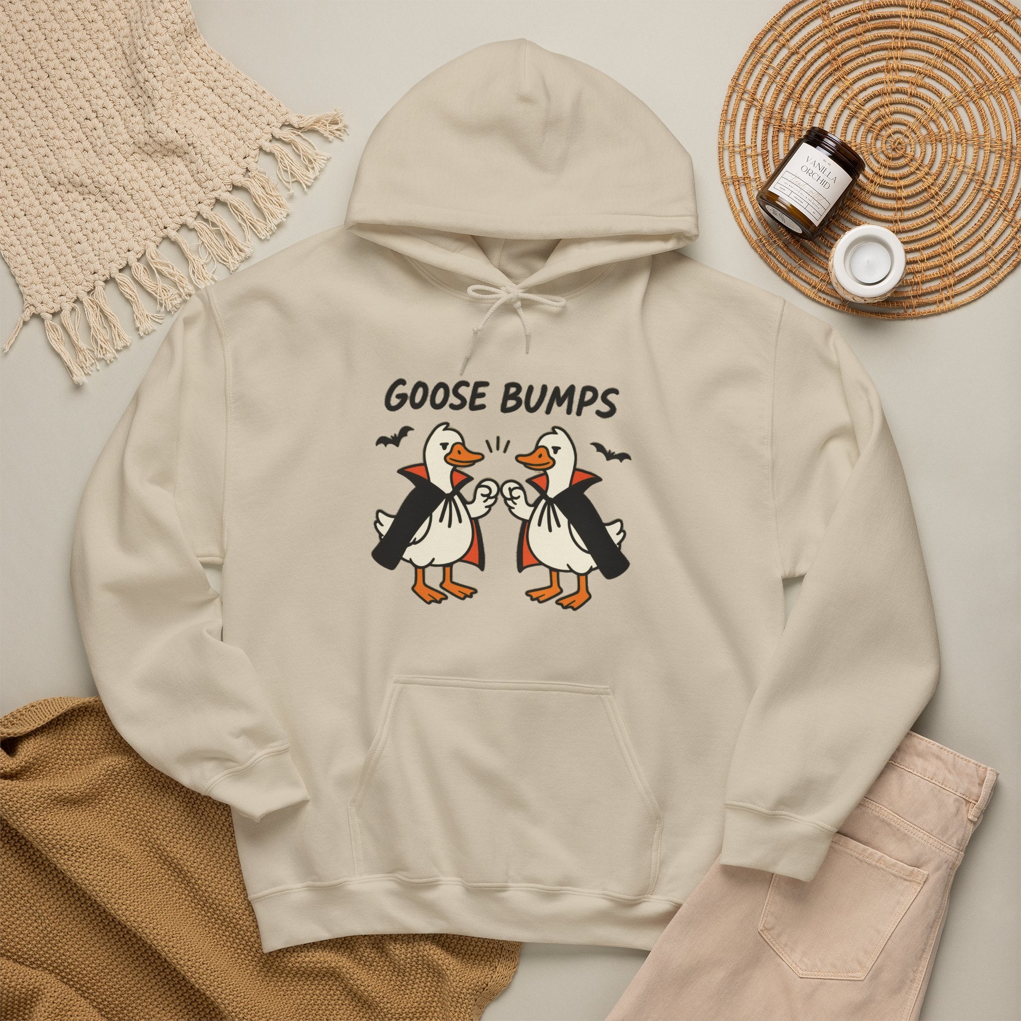 A sand-colored hoodie with a graphic of two penguins in black and white costumes with orange beaks, under the text 'GOOSE BUMPS'. The hoodie is styled in a flat-lay arrangement with a beige knit blanket, a brown knit sweater, light pink pants, and a small jar on a woven mat.