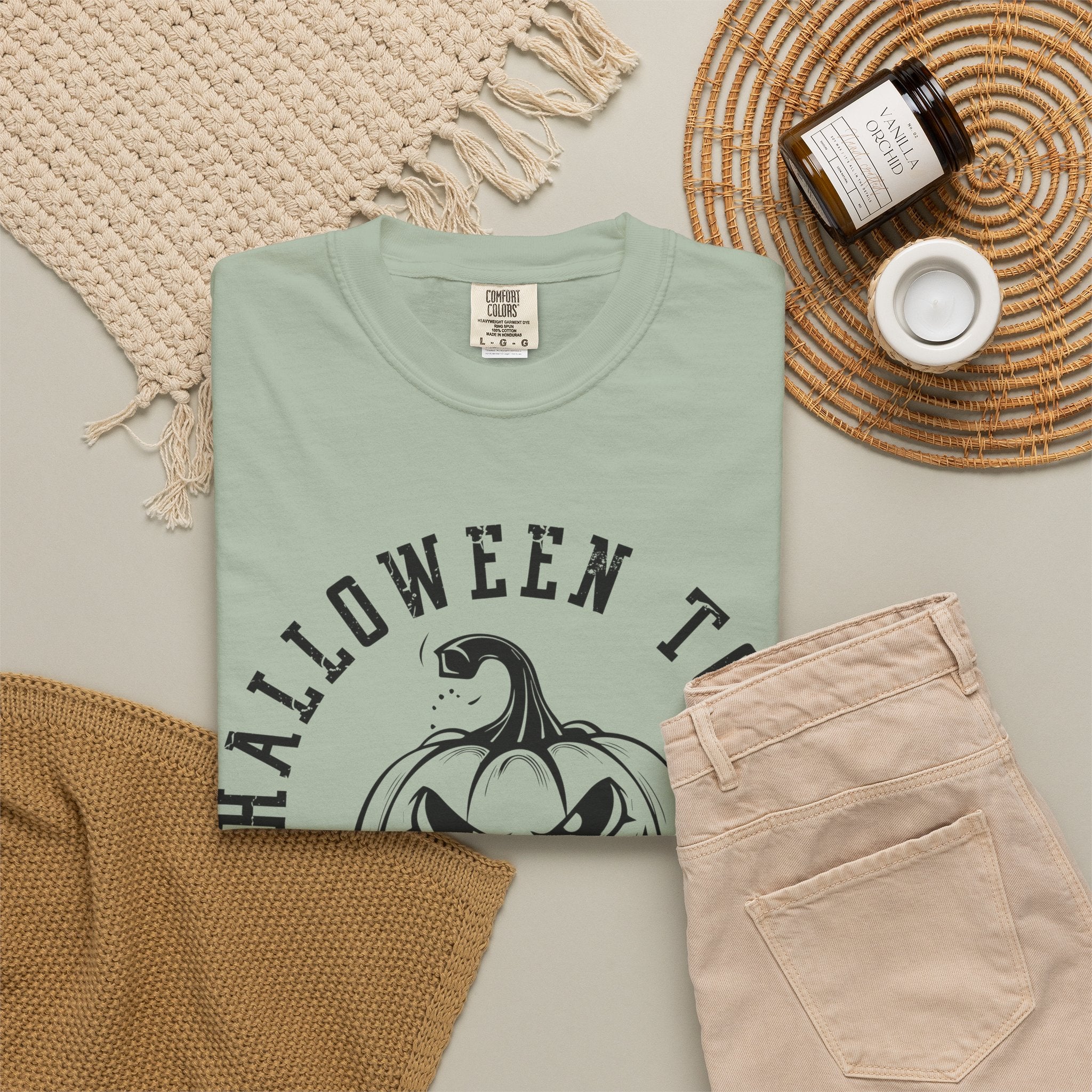 A folded bay colored-shirt with a black graphic of a pumpkin-headed creature and the text 'HALLOWEEN TH' is styled in a flat lay with beige pants, a woven blanket, a candle jar, and a small cup on a neutral background.