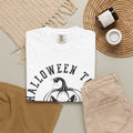 A folded white graphic T-shirt with a black Halloween-themed print reading 'HALLOWEEN TO' above a pumpkin illustration, styled in a flat lay with beige pants, woven textiles, a candle jar, and a small cup on a light neutral background.