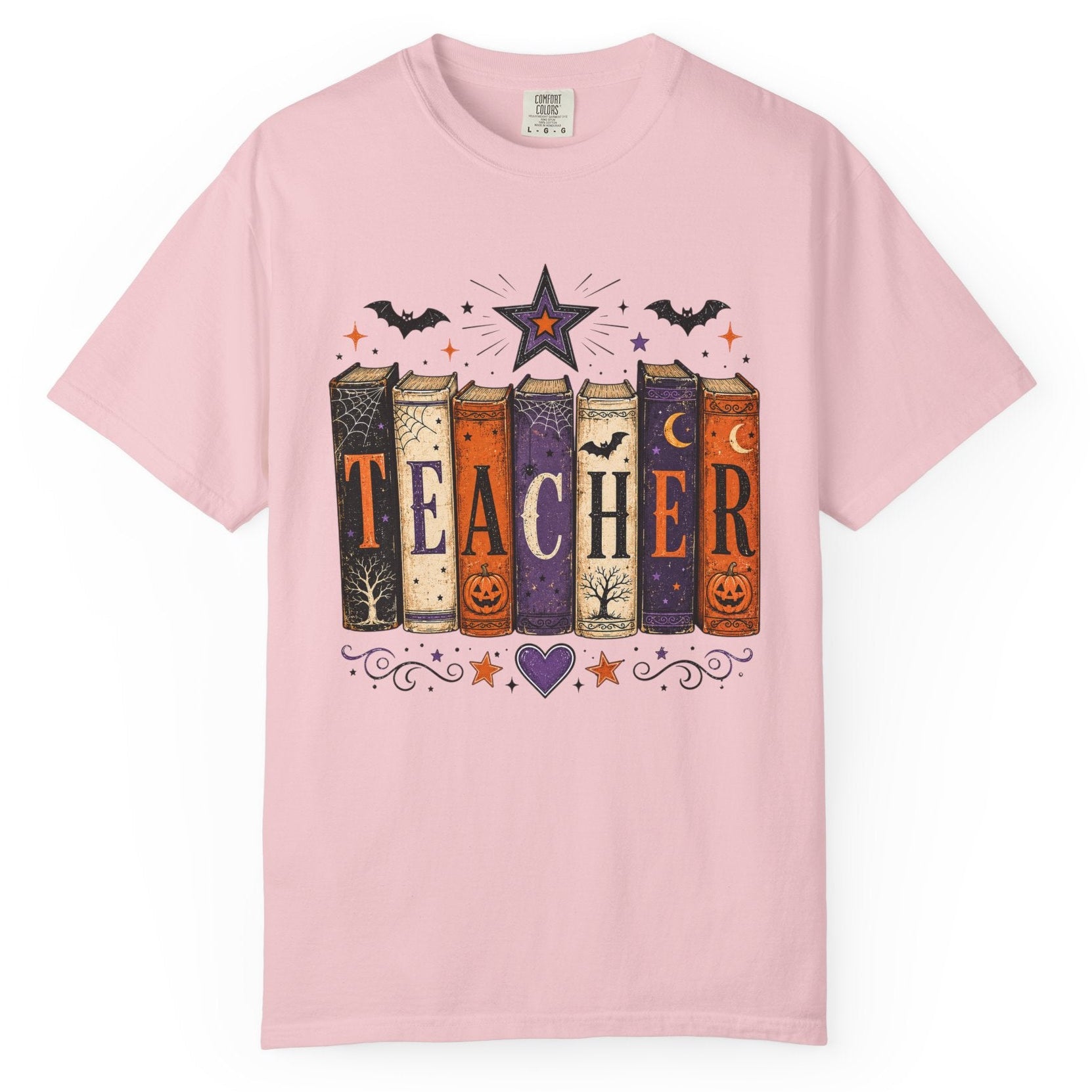 A light pink short-sleeve crewneck t-shirt featuring a Halloween-themed graphic of six books spelling 'TEACHER' with bats, stars, and pumpkins.