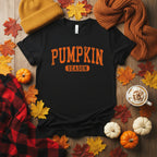 A black short-sleeve crewneck t-shirt with 'PUMPKIN SEASON' printed on the chest in distressed orange lettering, displayed with fall accessories
