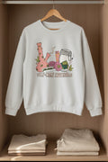 A sport gray crewneck sweatshirt featuring a colorful graphic of self-care and healing elements, including a pink bong, a jar, a slice of watermelon, and a heart, with the text 'SELF-CARE ESSENTIALS' printed below.