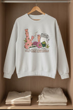 A sport gray crewneck sweatshirt featuring a colorful graphic of self-care and healing elements, including a pink bong, a jar, a slice of watermelon, and a heart, with the text 'SELF-CARE ESSENTIALS' printed below.