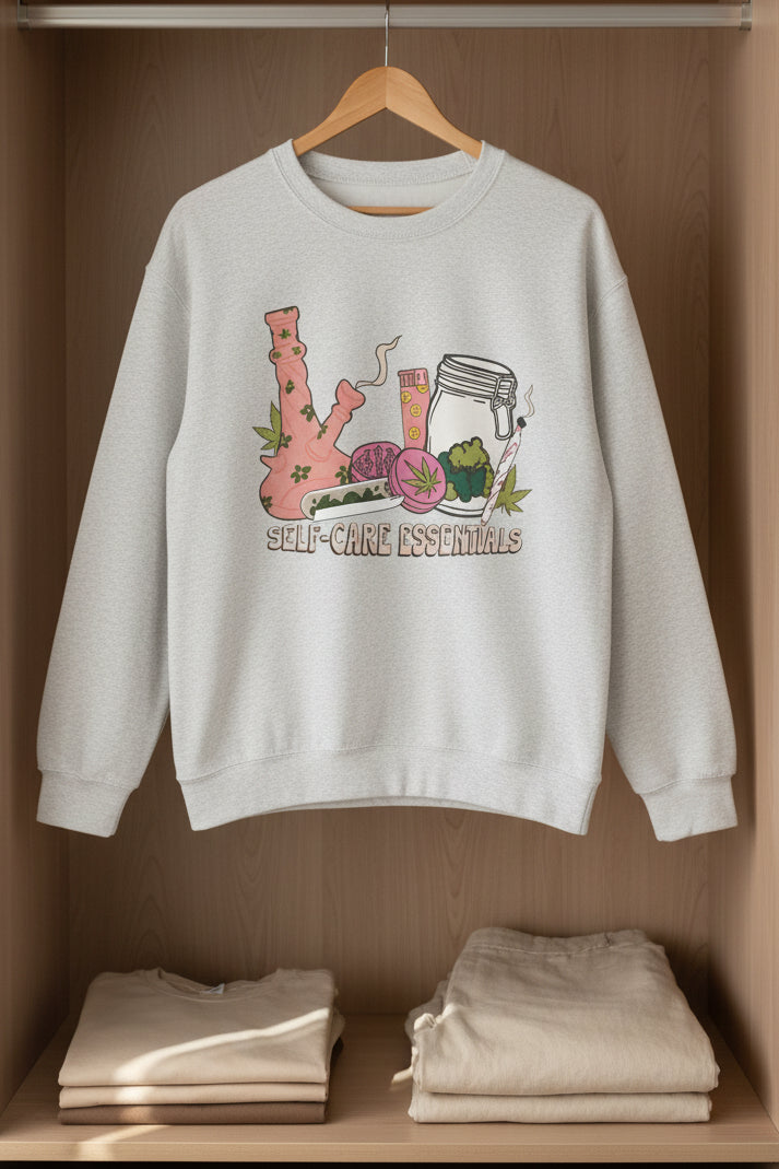 A sport gray crewneck sweatshirt featuring a colorful graphic of self-care and healing elements, including a pink bong, a jar, a slice of watermelon, and a heart, with the text 'SELF-CARE ESSENTIALS' printed below.