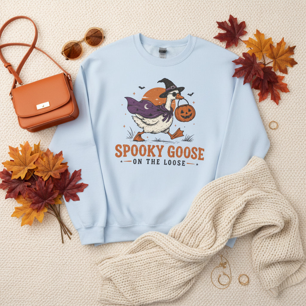A light blue crewneck sweatshirt featuring a cartoon graphic of a goose dressed as a witch holding a jack-o'-lantern, with the text 'SPOOKY GOOSE ON THE LOOSE' printed below.