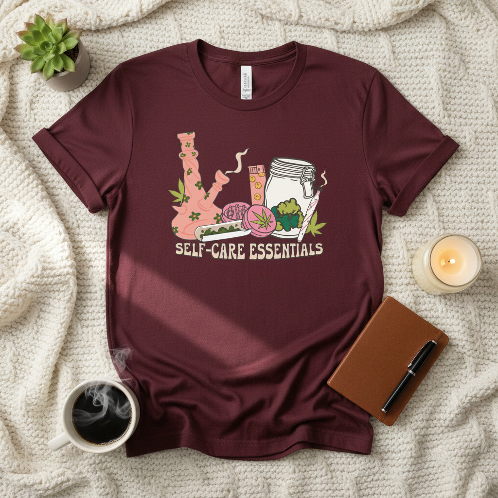 A maroon short-sleeve t-shirt with a colorful graphic on the front showing a pink bong, a glass jar, a pink cup, and a green leaf, with the text 'SELF-CARE ESSENTIALS' printed below.