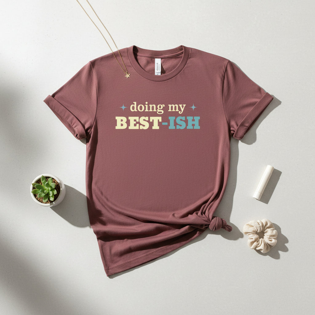 A short-sleeve crewneck t-shirt in a dusty rose or mauve color, featuring the text 'doing my BEST-ISH' printed across the chest in cream, yellow, and light blue. The t-shirt is displayed flat against a solid white background.
