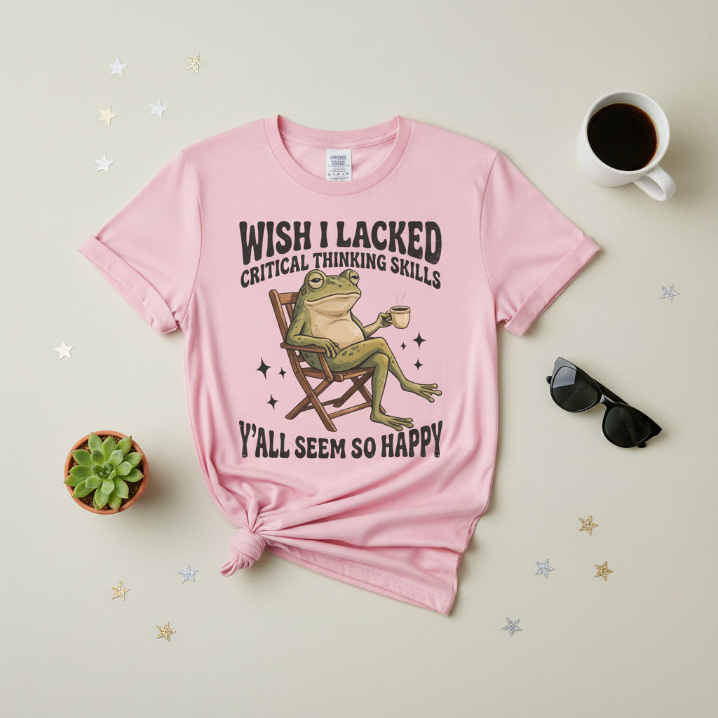 A light pink short-sleeve t-shirt with a graphic of a green frog sitting in a wooden chair and holding a mug, with the text 'WISH I LACKED CRITICAL THINKING SKILLS' above and 'Y'ALL SEEM SO HAPPY' below.