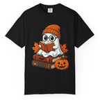A black short-sleeve crewneck t-shirt featuring a cartoon graphic of a ghost wearing glasses and an orange knit beanie, sitting on books and reading an orange book, with autumn leaves and a jack-o'-lantern pumpkin.
