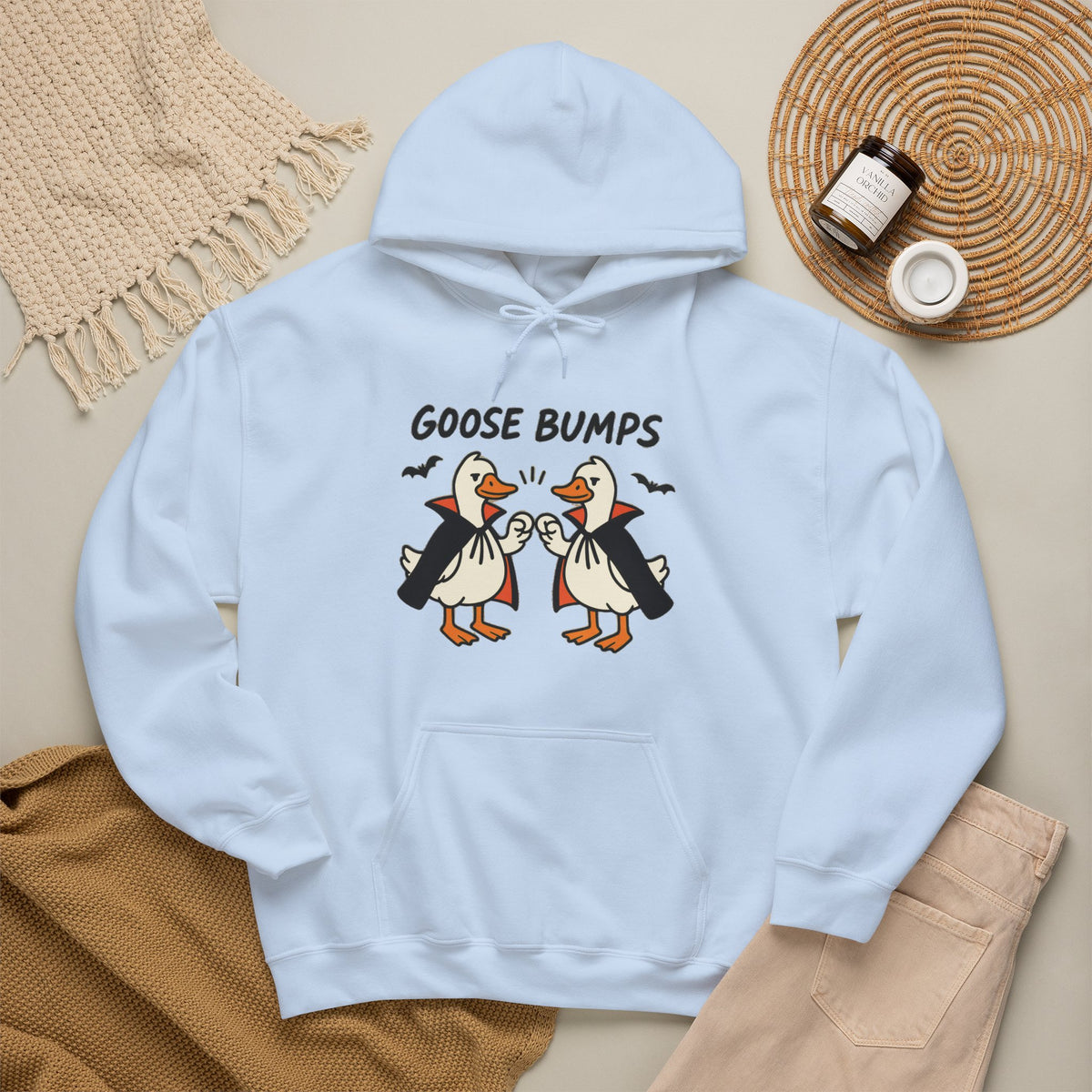 A light blue hoodie with a graphic of two penguins in black and white costumes with orange beaks, under the text 'GOOSE BUMPS'. The hoodie is styled in a flat-lay arrangement with a beige knit blanket, a brown knit sweater, light pink pants, and a small jar on a woven mat.