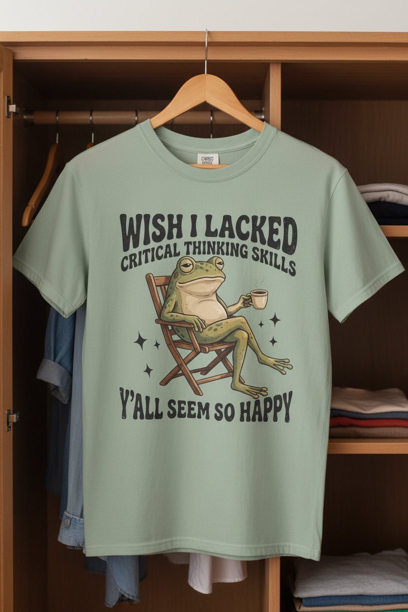 A sage green short-sleeve t-shirt with a graphic of a frog sitting in a director's chair and the text 'WISH I LACKED CRITICAL THINKING SKILLS' and 'Y'ALL SEEM SO HAPPY'.