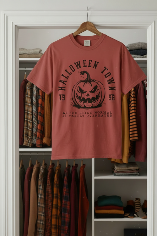 A crimson-colored short-sleeve t-shirt with a black graphic of a jack-o'-lantern and the text 'HALLOWEEN TOWN' and 'WHERE BEING NORMAL IS VASTLY OVERNIGHTED', hanging on a wooden hanger in front of an open white closet door with folded clothes.