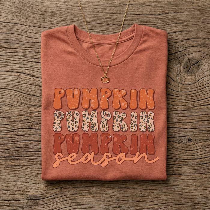A folded heather clay t-shirt with a 'Pumpkin Pumpkin Pumpkin season' graphic print, styled on a rustic wood background with a gold necklace.