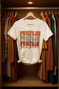 An soft cream short-sleeve t-shirt with a retro 'Pumpkin Season' graphic, styled in a flat-lay on a dark wood background with a coffee mug, book, and cutlery.