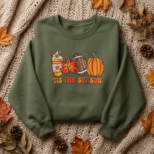 An military green crewneck sweatshirt with a festive autumn graphic featuring a hot chocolate cup, maple leaf, football, pumpkin, and the text 'TIS THE SEASON'. The sweatshirt is displayed in a flat-lay arrangement on a beige knit blanket, surrounded by decorative fall leaves, pinecones, and dried botanicals.