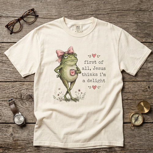 A cream-colored graphic t-shirt laid flat on a rustic wooden surface, surrounded by lifestyle props including a pair of glasses, a watch, and a pocket compass. The t-shirt features a cartoon illustration of a green frog wearing a pink bow and a small pink cross, next to the text 'first of all, Jesus thinks i'm a delight' in a vintage typewriter font.