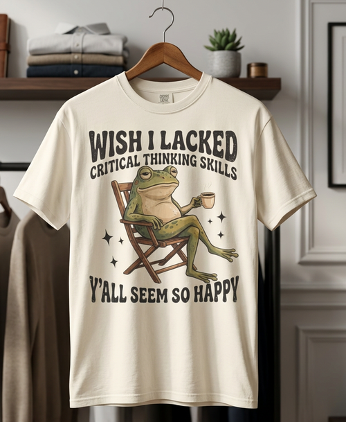 A cream-colored graphic t-shirt hanging on a wooden hanger in a styled room setting. The shirt features a cartoon frog sitting in a director's chair holding a mug, with the text 'WISH I LACKED CRITICAL THINKING SKILLS' above and 'Y'ALL SEEM SO HAPPY' below.