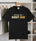 A black short-sleeve t-shirt hanging on a wooden hanger against a blurred closet background. The shirt features the text 'doing my BEST-ISH' printed on the chest, with 'doing my' and 'BEST-ISH' in yellow and white respectively.