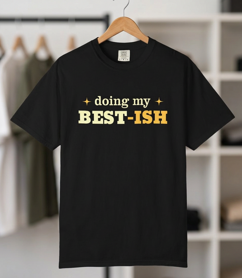 A black short-sleeve t-shirt hanging on a wooden hanger against a blurred closet background. The shirt features the text 'doing my BEST-ISH' printed on the chest, with 'doing my' and 'BEST-ISH' in yellow and white respectively.