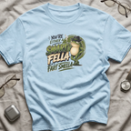 A light blue graphic t-shirt laid flat on a beige fabric background, surrounded by lifestyle props including eyeglasses, a watch, and a wireless earbud case. The shirt features a graphic of a green frog wearing a black hat and the text 'YOU'RE EITHER SMART FELLA  OR A FART SMELLA'.