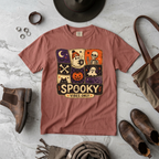 A dusty rose short-sleeve graphic t-shirt laid flat on a light gray marble surface, surrounded by autumn and Halloween styling props including brown leather boots, a hat, a scarf, rings, and a tote bag. The t-shirt features a square patchwork graphic with ghosts, a skeleton, a jack-o'-lantern, and the text 'SPOOKY VIBES ONLY.