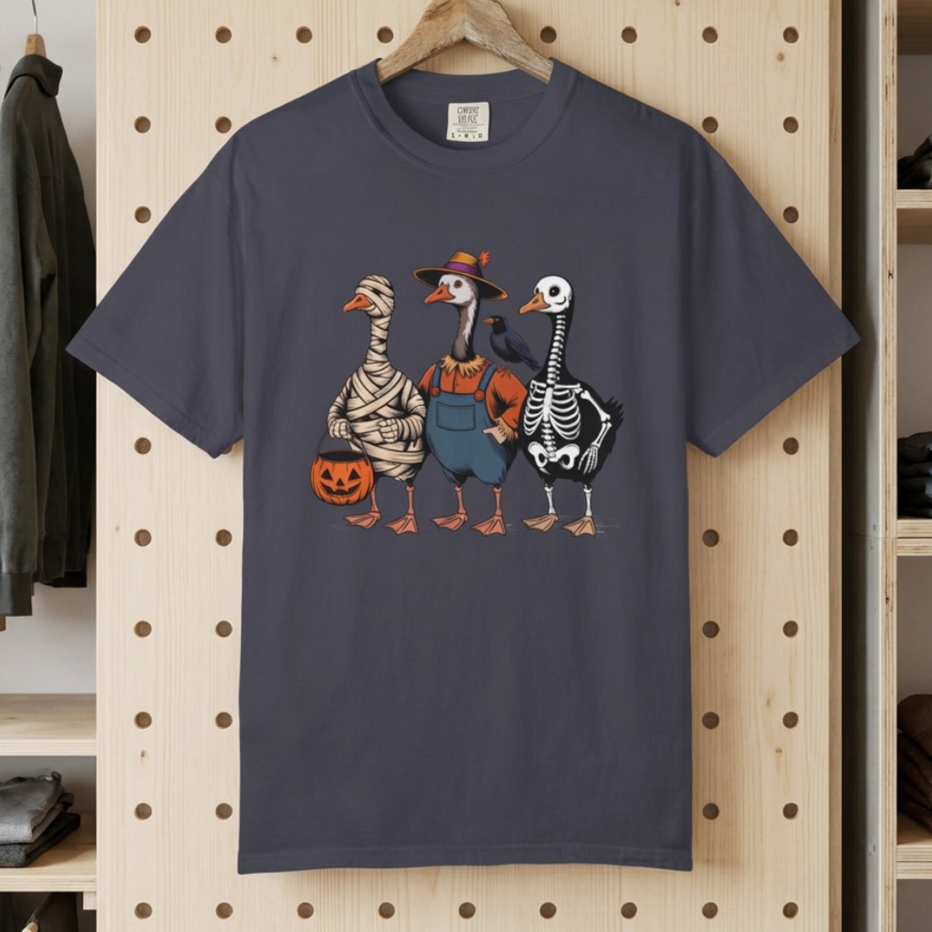 A dark gray crewneck t-shirt hanging on a wooden hanger against a light wood pegboard wall. The shirt features a graphic of three geese dressed as mummies, a in blue o goose overalls, and a goose dressed as a skeleton, each holding a jack-o'-lantern.